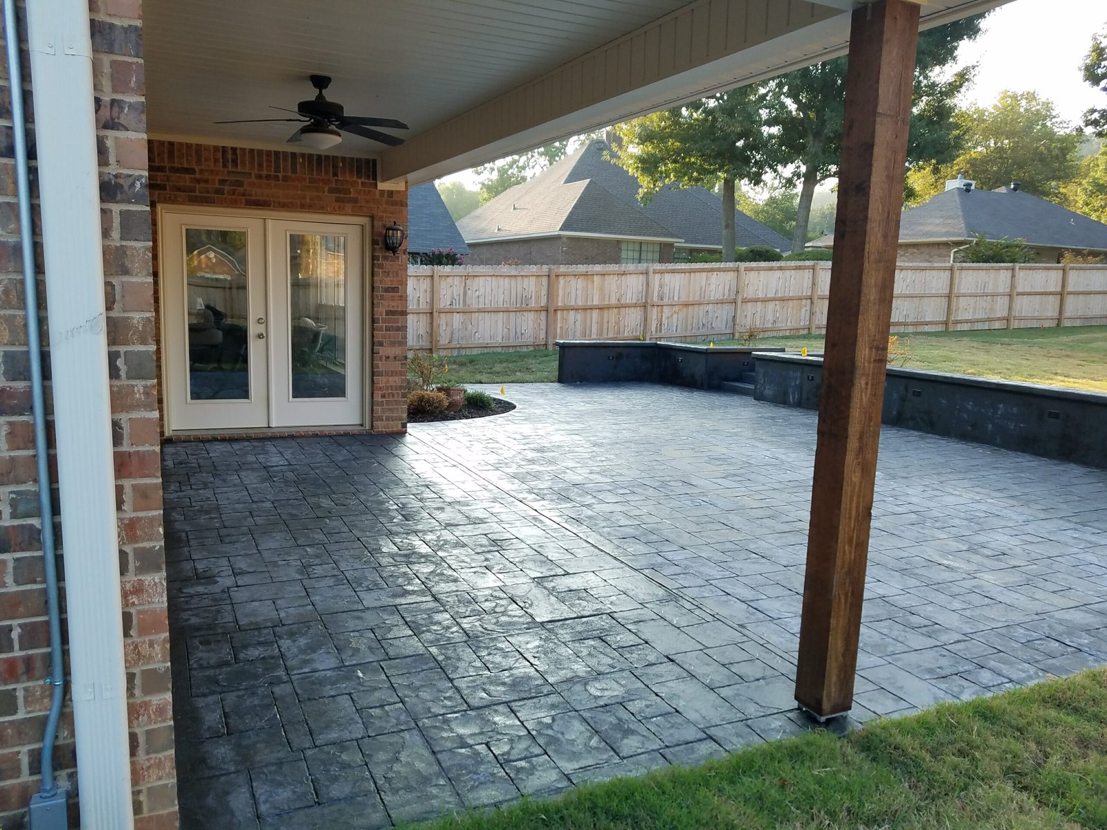 A covered patio with a stamped concrete floor, wooden support post, ceiling fan, and double glass doors opens to a fenced backyard with grass, lush trees in the background, and a small garden bed.