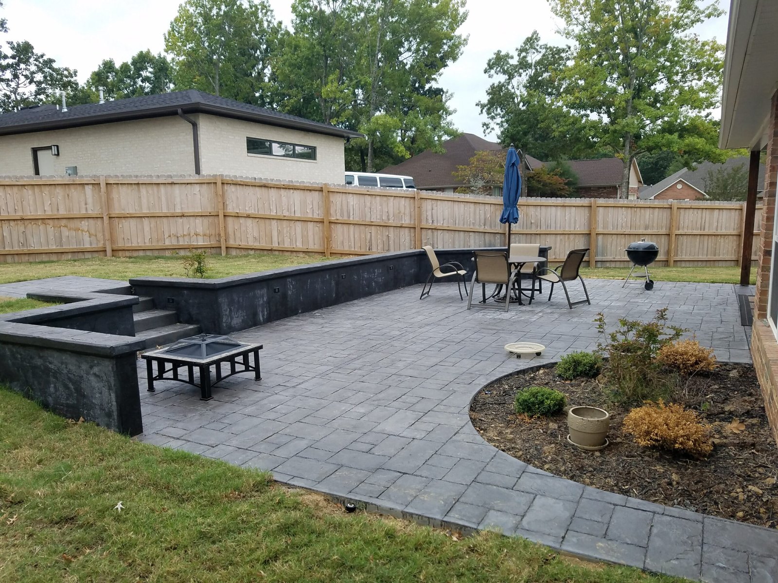 A backyard patio with gray stone pavers, built-in black benches, a cozy seating area with a table, four chairs, a blue umbrella, grill, and a lush curved garden bed with plants—all surrounded by a wooden privacy fence.