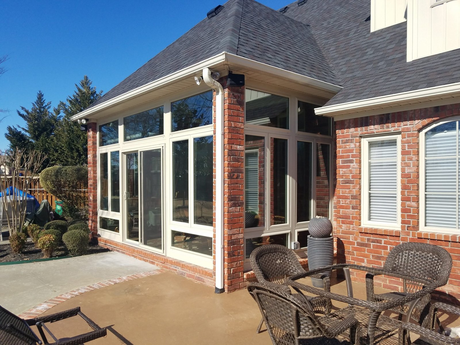 A sunroom with large windows and a brick exterior connects to a patio with wicker chairs and a table. Manicured bushes, shade trees, and clear blue sky create a relaxing outdoor retreat.
