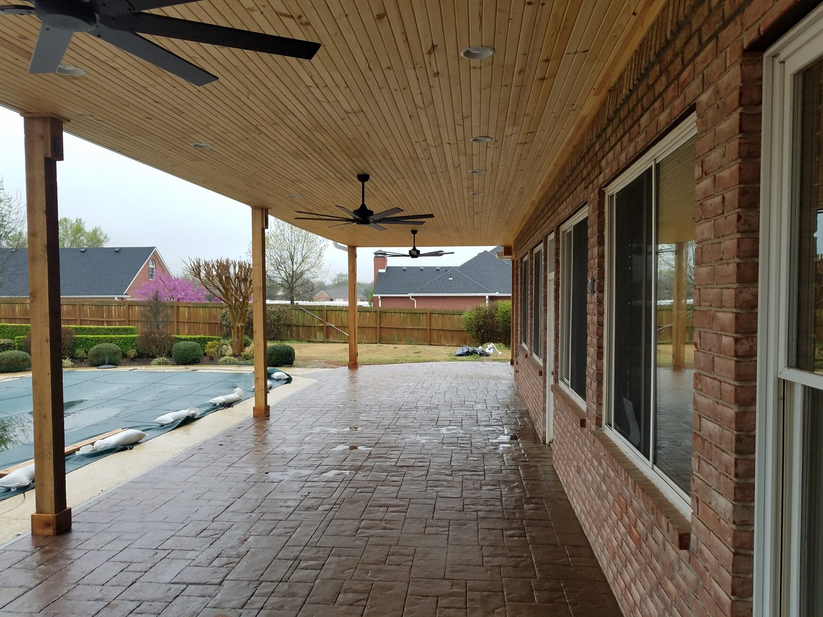 Covered backyard patio with a stamped concrete floor, wooden ceiling, ceiling fans, brick wall, and large windows. This inviting space overlooks a fenced yard with a covered pool and well-trimmed bushes for private relaxation.
