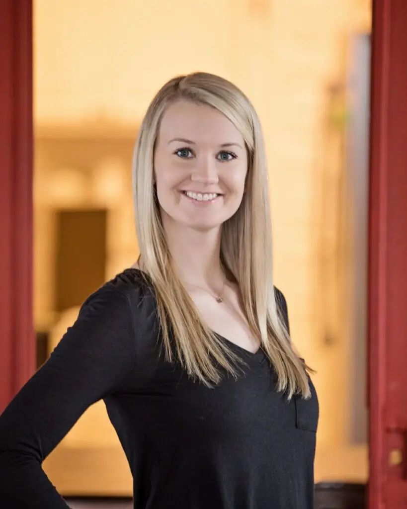 A woman with straight blonde hair wearing a black long-sleeve shirt stands indoors, smiling at the camera. The background is warmly lit with red modern framing elements.