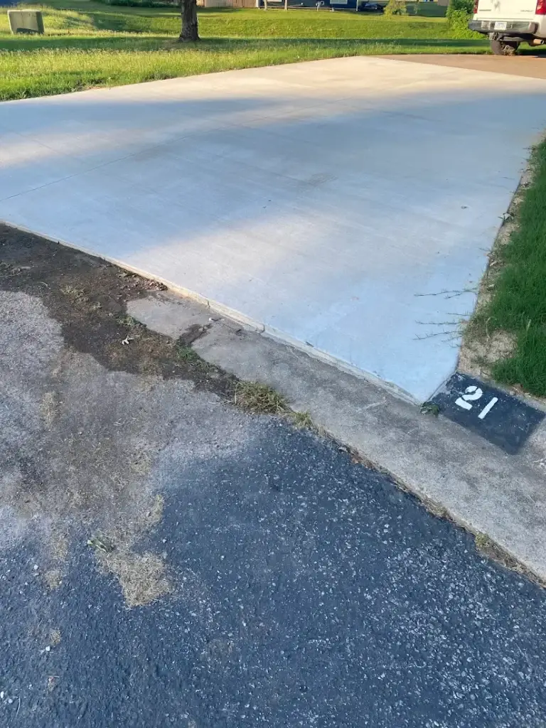 A newly paved concrete driveway adjoins an older asphalt surface, with the number 21 painted on a small black square near the concrete’s edge. Grass and a well-kept lawn provide a green backdrop.