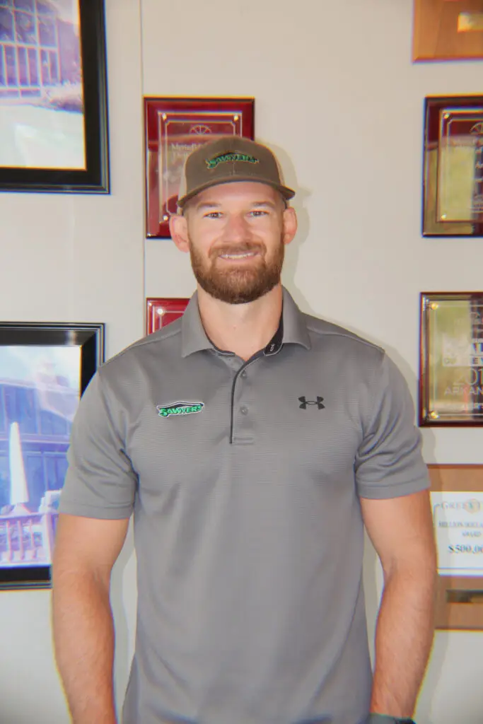 A man with a beard and a gray cap smiles at the camera. He is wearing a gray collared shirt featuring an embroidered green and black logo. Framed certificates and sustainability awards are displayed on the wall behind him.