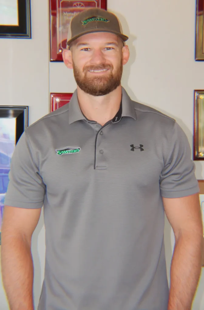A man with a beard and a gray cap smiles at the camera. He is wearing a gray collared shirt featuring an embroidered green and black logo. Framed certificates and sustainability awards are displayed on the wall behind him.