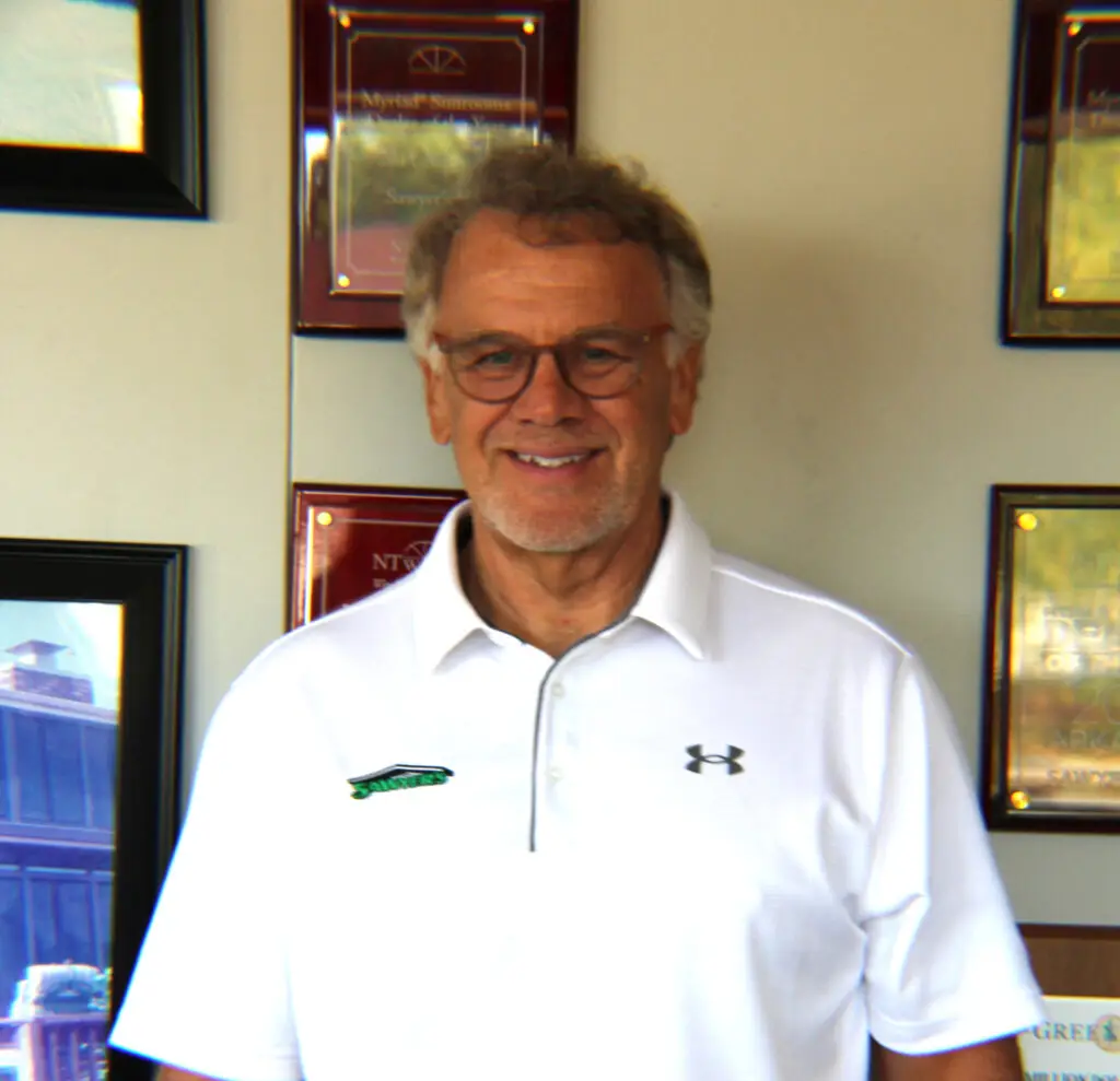 A smiling older man with gray hair and glasses wears a white Under Armour polo shirt. He stands indoors in front of a wall decorated with framed awards and certificates, highlighting his achievements.