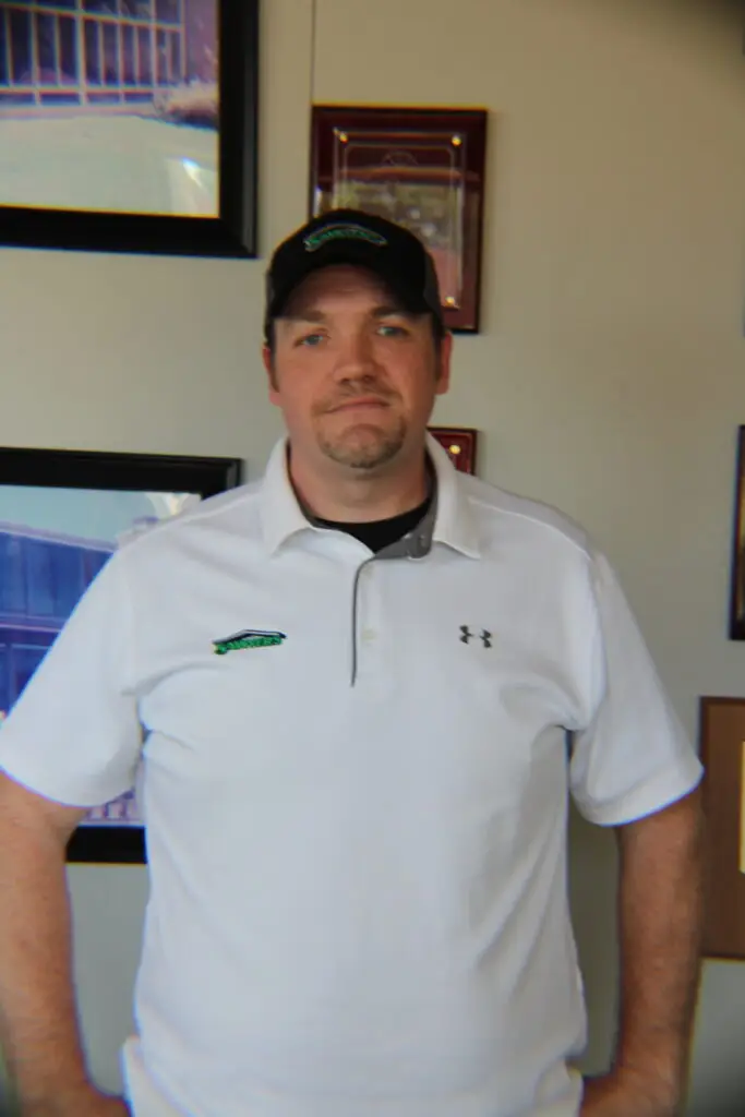 A man wearing a white Under Armour polo shirt and a black cap stands indoors with his hands on his hips, in front of a gallery wall decorated with framed pictures and plaques.