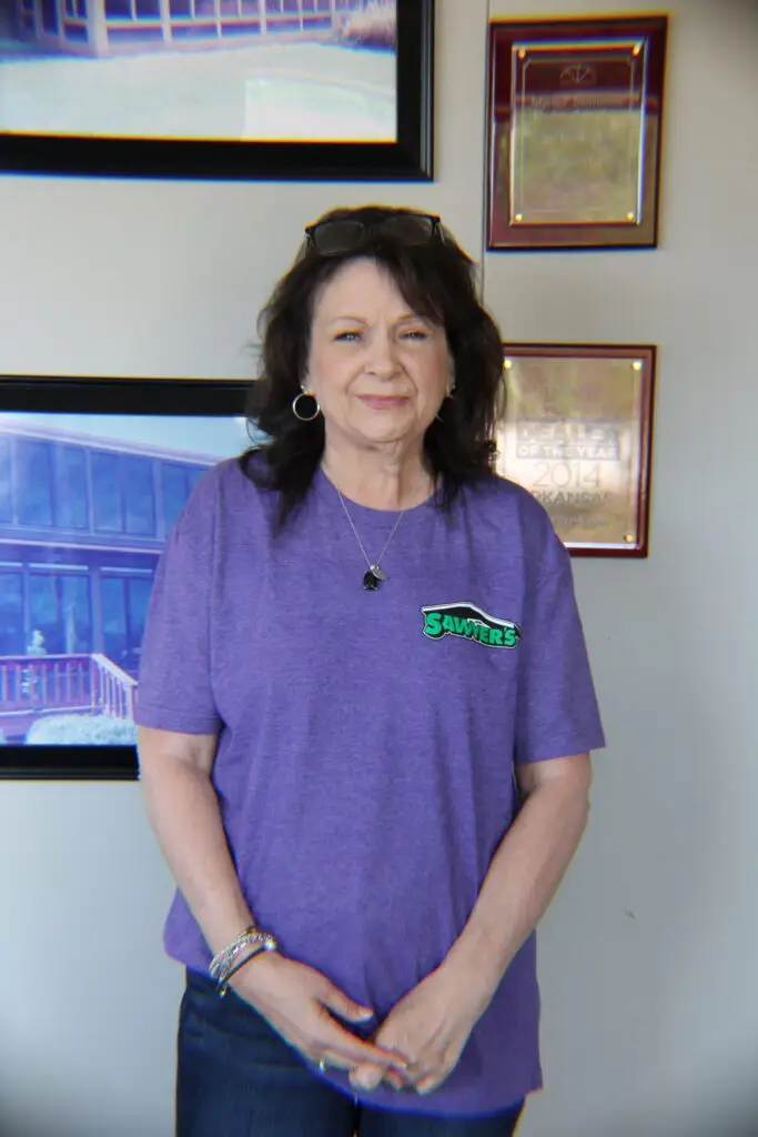 A woman with dark hair wearing a purple Sawyers t-shirt and bracelets stands indoors, smiling in front of a wall decorated with framed photos and awards, celebrating the Sawyers brand.