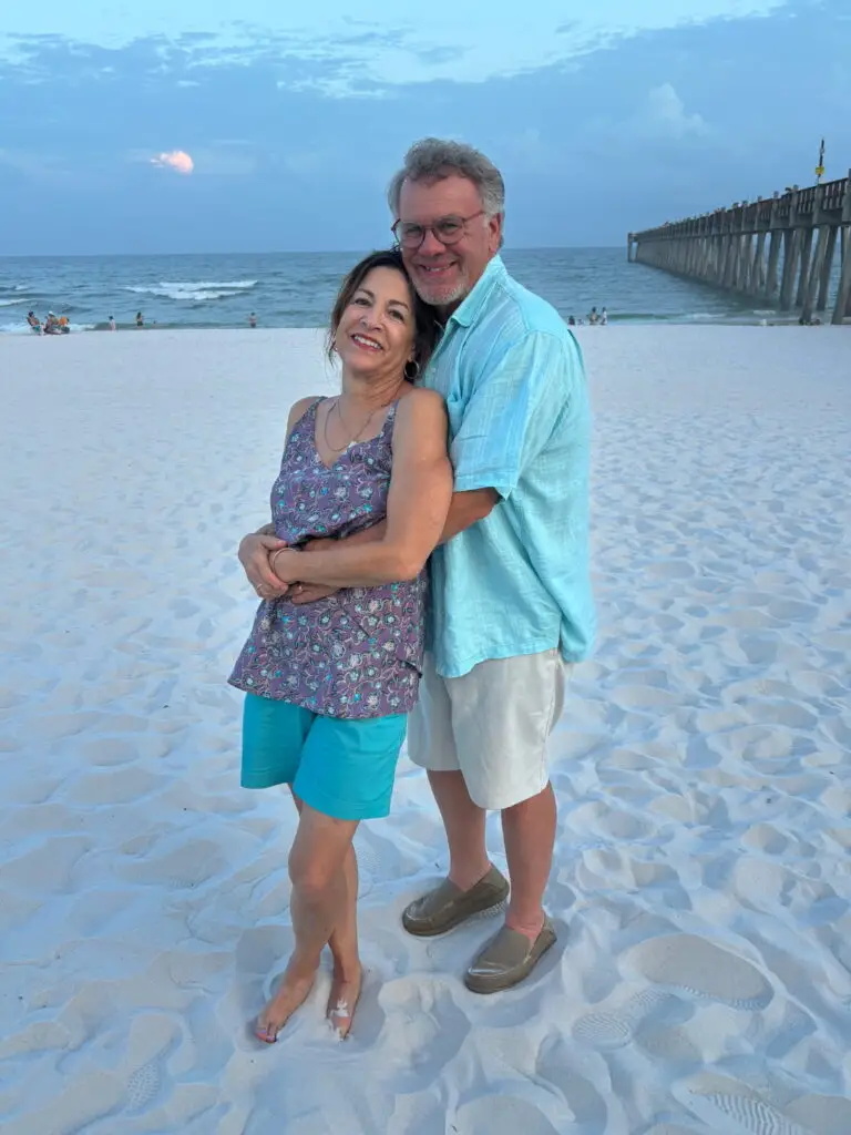 A smiling couple embraces on a sandy beach near the ocean, with a scenic pier extending into the water and people in the background. The sky is mostly clear with some clouds, creating a romantic coastal atmosphere.