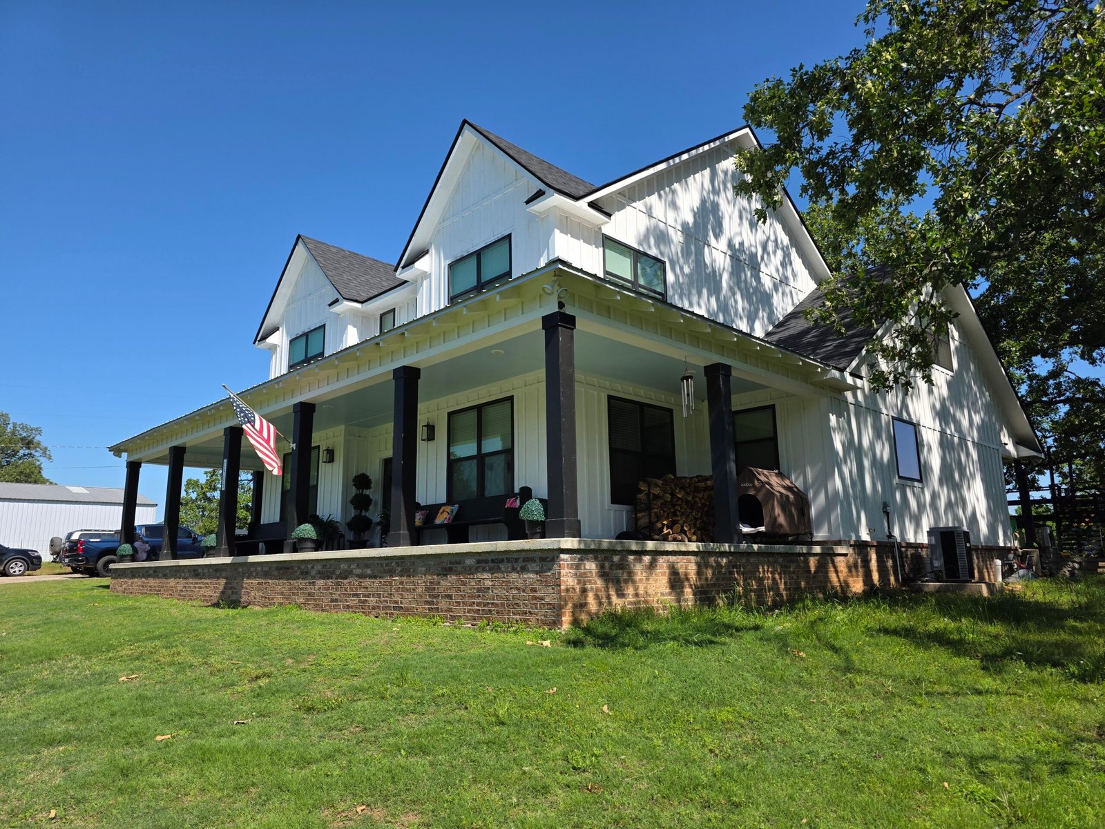 A modern white two-story house with black trim and a large wraparound porch, featuring a U.S. flag, porch chairs, cozy firewood, and surrounded by a lush grassy yard under a clear blue sky.