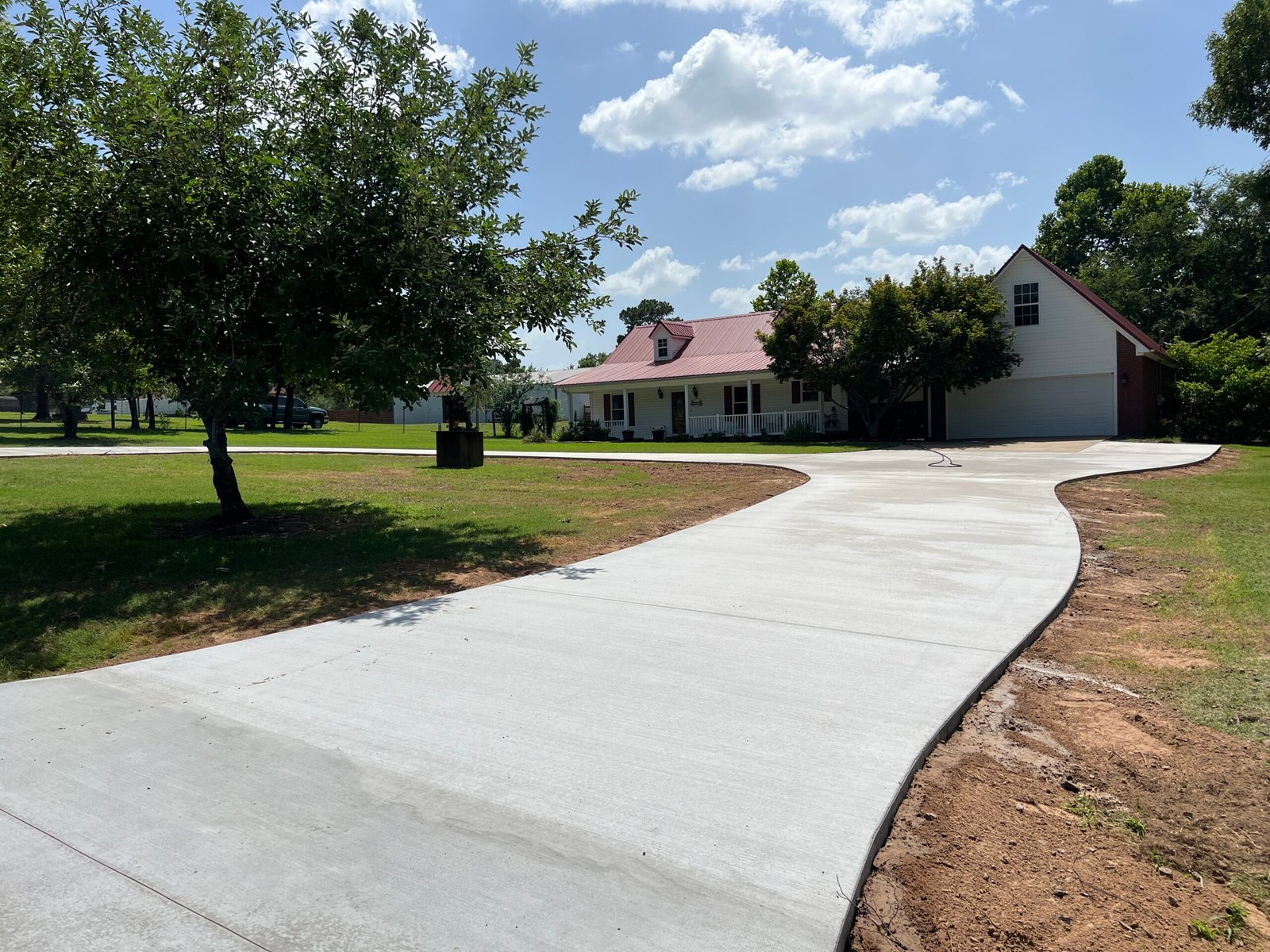 A long, curved concrete driveway leads to a charming white house with a red roof and attached garage, surrounded by green grass, trees, and a partly cloudy sky.