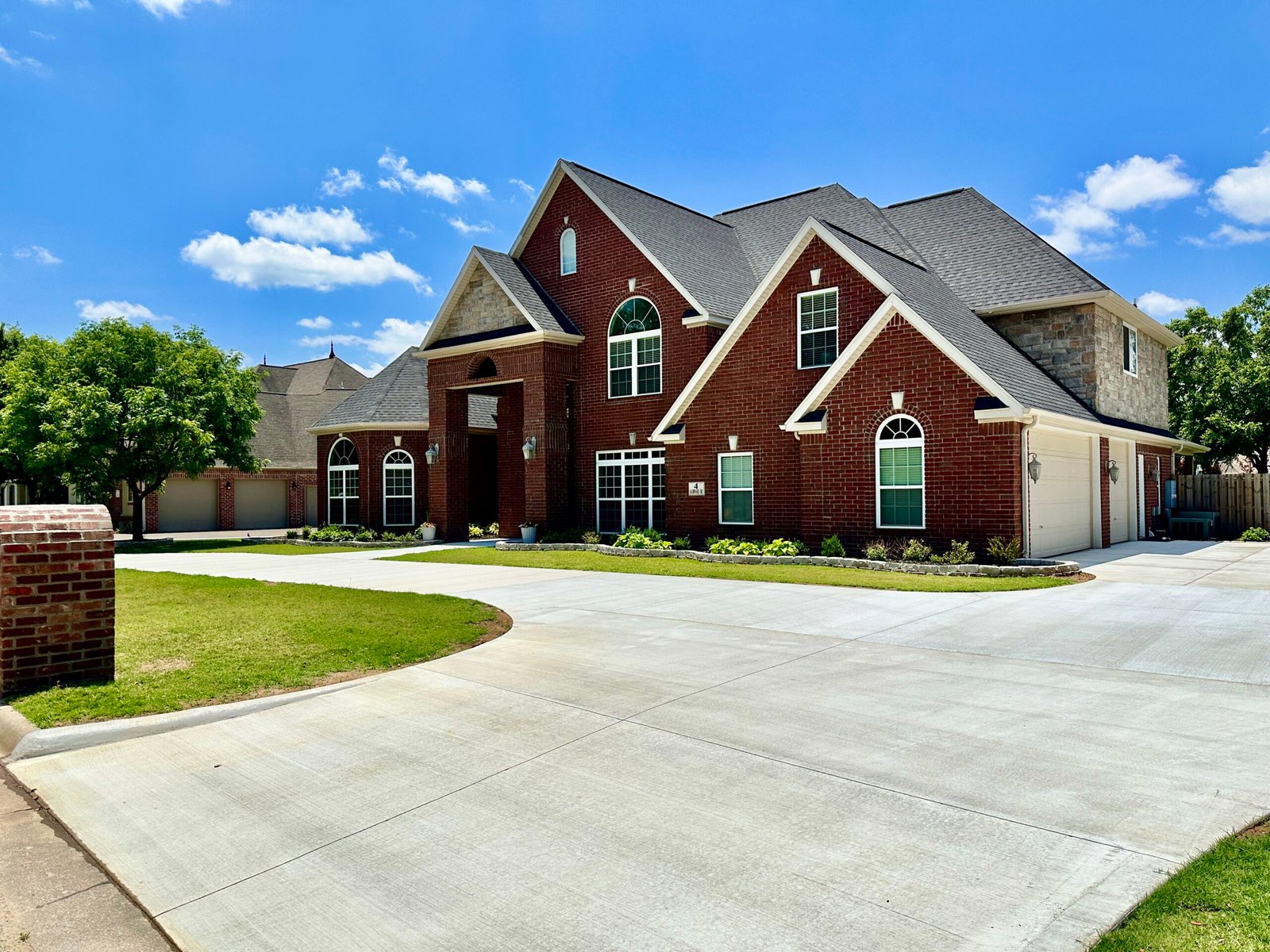 A large two-story brick house with white trim, multiple gables, and arched windows sits under a clear blue sky with scattered clouds. The property features a spacious driveway, green lawn, trees, and elegant landscaping.