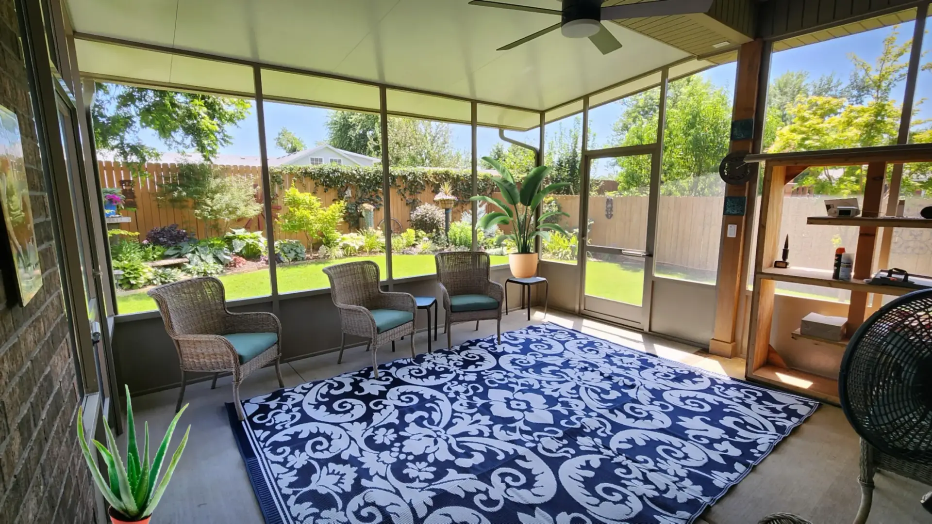A bright sunroom with wicker chairs, a blue-and-white patterned rug, ceiling fan, and expansive windows overlooking a lush green backyard garden filled with trees and plants—perfect for relaxing in natural light.