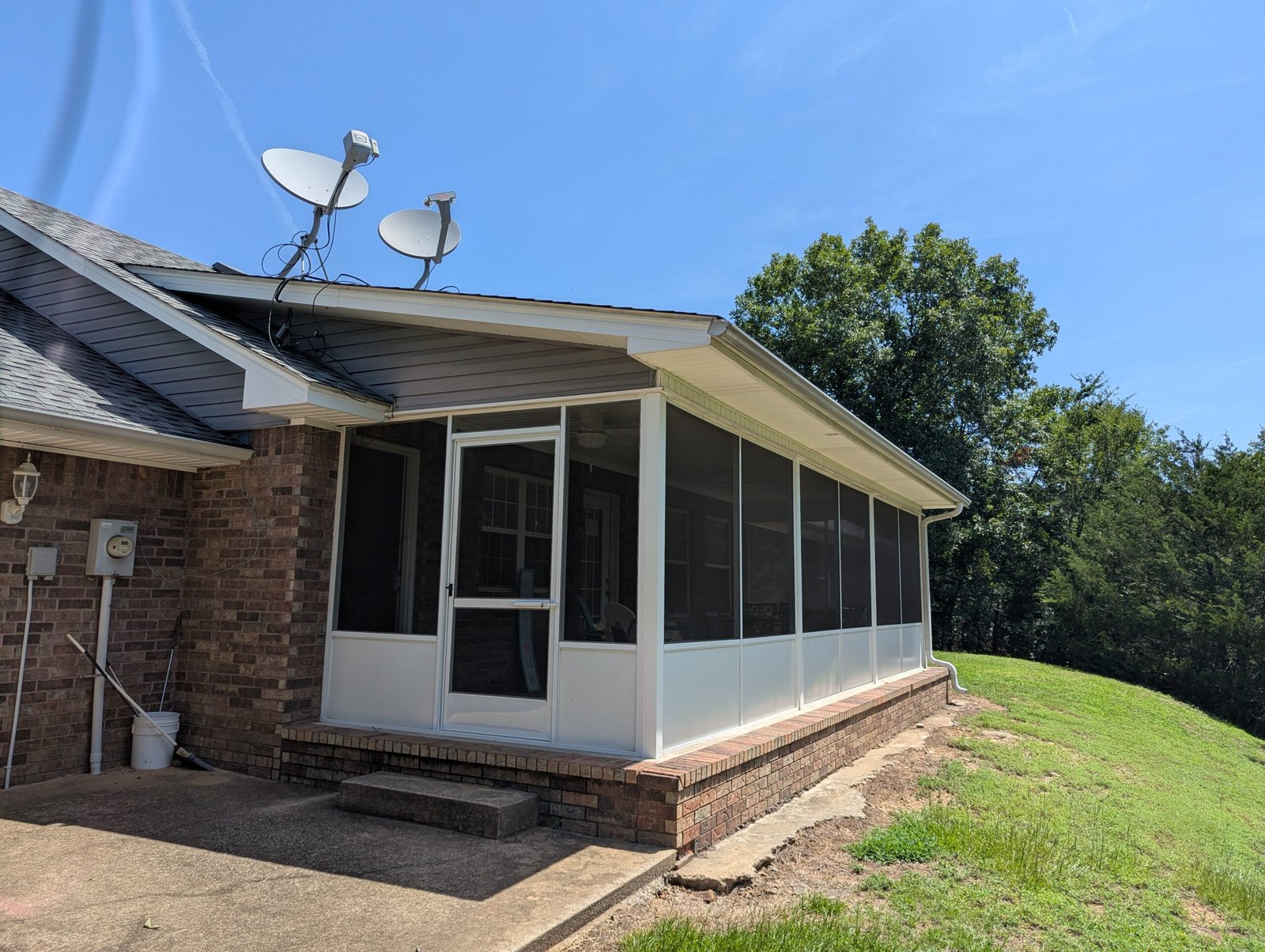 A brick house with a screened-in porch, two satellite dishes on the roof, and a grassy yard sloping down beside the porch under a clear blue sky offers modern connectivity in a peaceful setting.