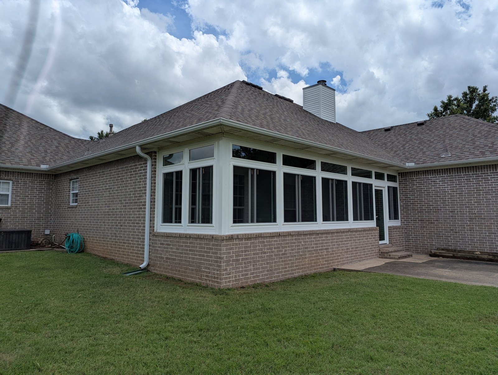A brick house with a sunroom featuring many windows, a sloped shingle roof, lush green lawn, hose reel, and partly cloudy sky in the background.