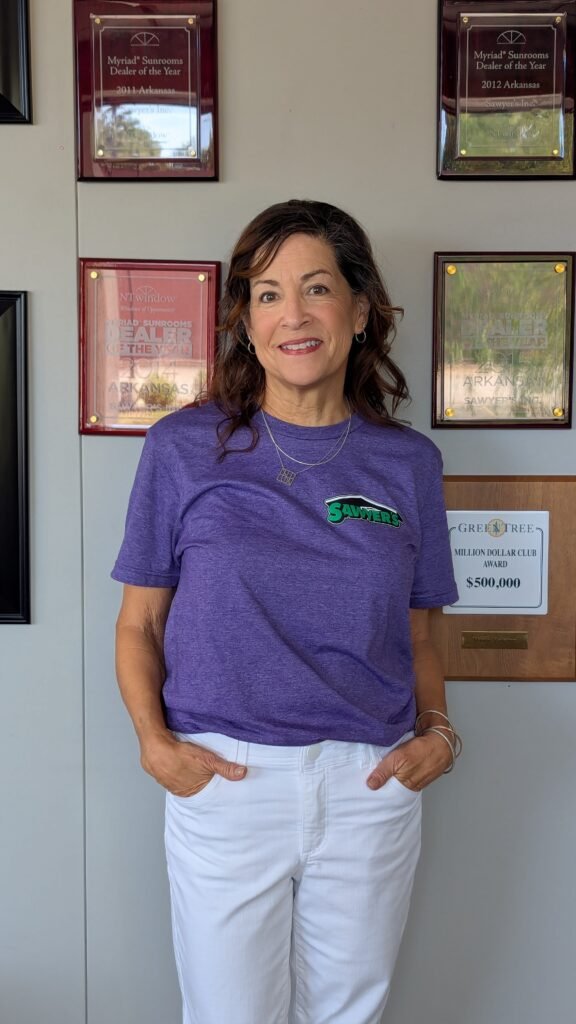 A woman with wavy brown hair, wearing a purple T-shirt and white pants, stands smiling in front of a wall showcasing several achievement plaques and awards.