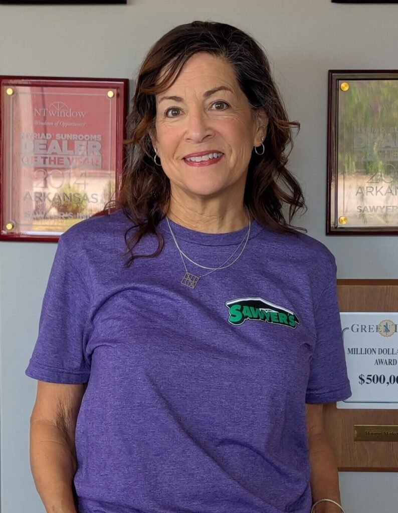 A woman with wavy brown hair, wearing a purple T-shirt and white pants, stands smiling in front of a wall showcasing several achievement plaques and awards.