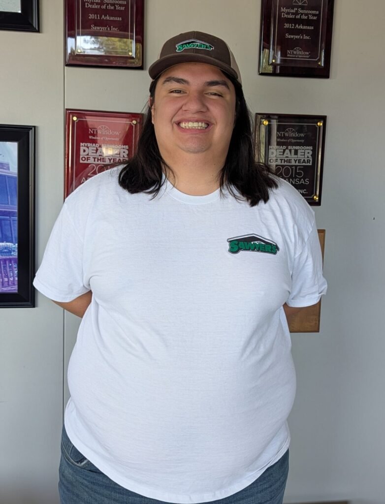 A person wearing a white t-shirt and brown cap stands indoors, smiling at the camera. Behind them, several award plaques and framed photos decorate the wall, highlighting achievements.
