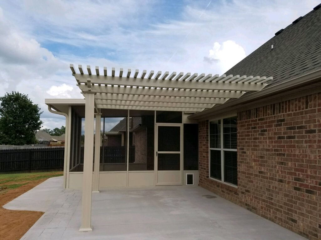 A white pergola extends over a concrete patio attached to a brick house with a screened porch door and windows. The outdoor space overlooks grass and a fenced backyard beneath a partly cloudy sky.
