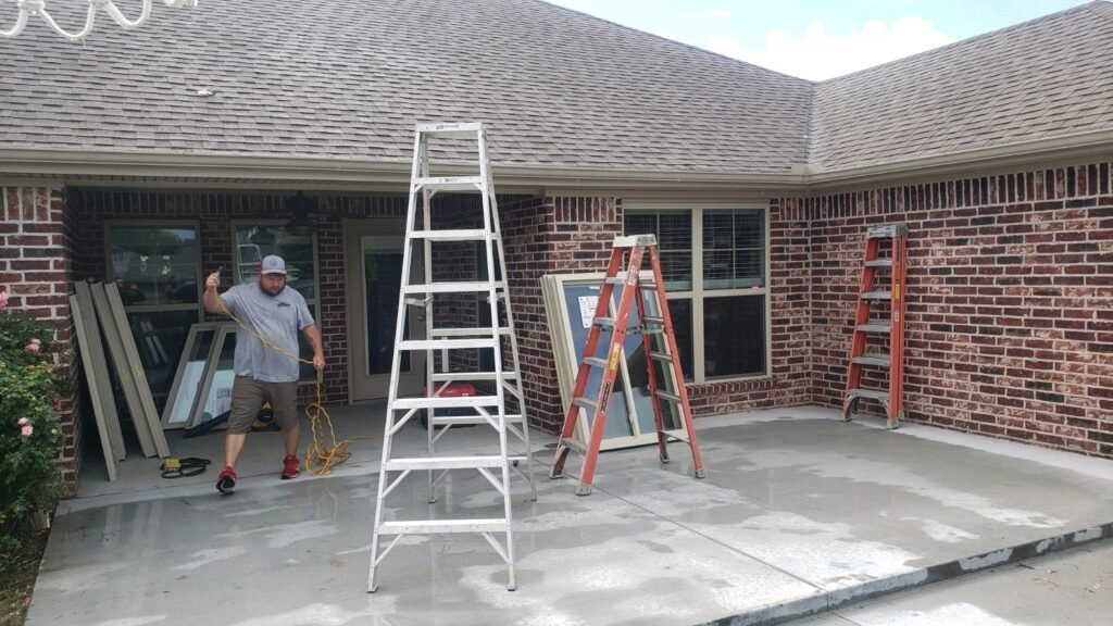 A man in a gray shirt and hat stands on a wet concrete patio near a brick house, holding a yellow cord. Three ladders and several window frames rest nearby. The house features large windows and a sloped roof, adding character to the scene.