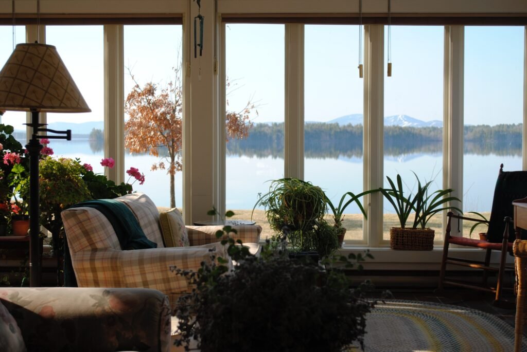 A sunlit living room with cozy chairs, lush houseplants, and large windows overlooking a calm lake, vibrant autumn trees, and distant mountains under a clear sky.