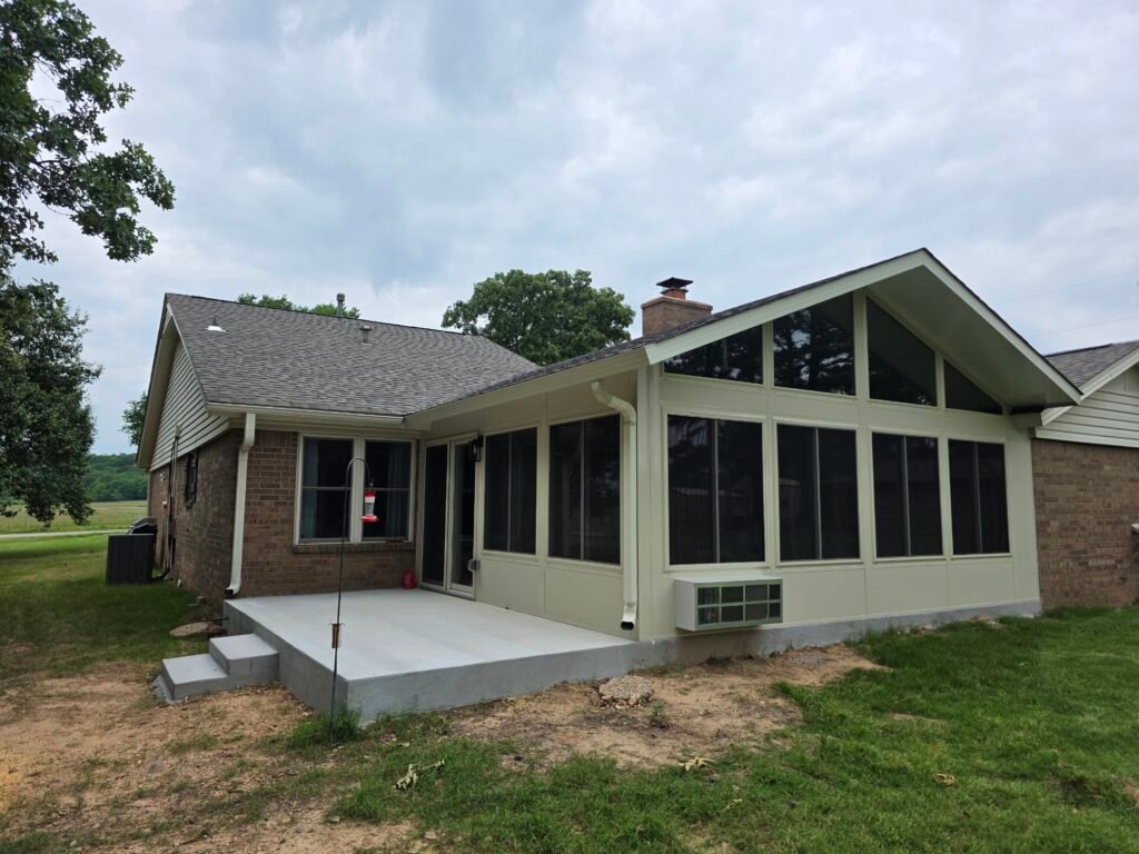 Enjoy the Arkansas Summer Without Hiding From It A brick house with a covered back porch enclosed by large screened windows, a concrete patio, and an air conditioning unit; the yard has grass and some bare patches, with trees and open field in the background.