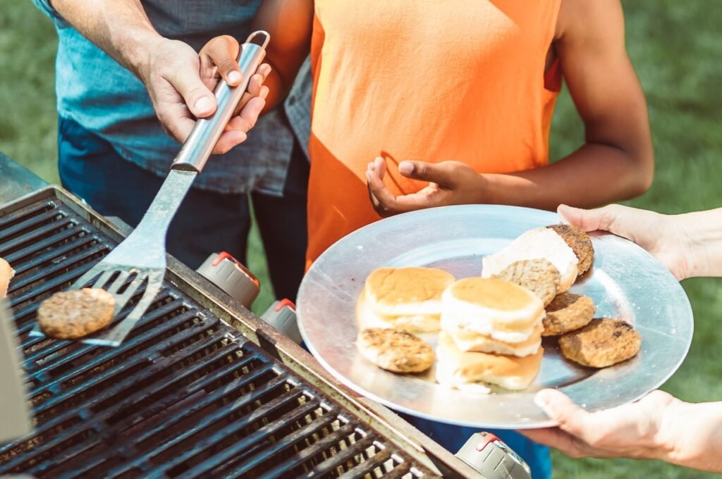 Three people prepare burgers at an outdoor grill. One person holds a spatula with a cooked patty, while another holds a metal plate with buns and burger patties. The scene is set on a sunny day.