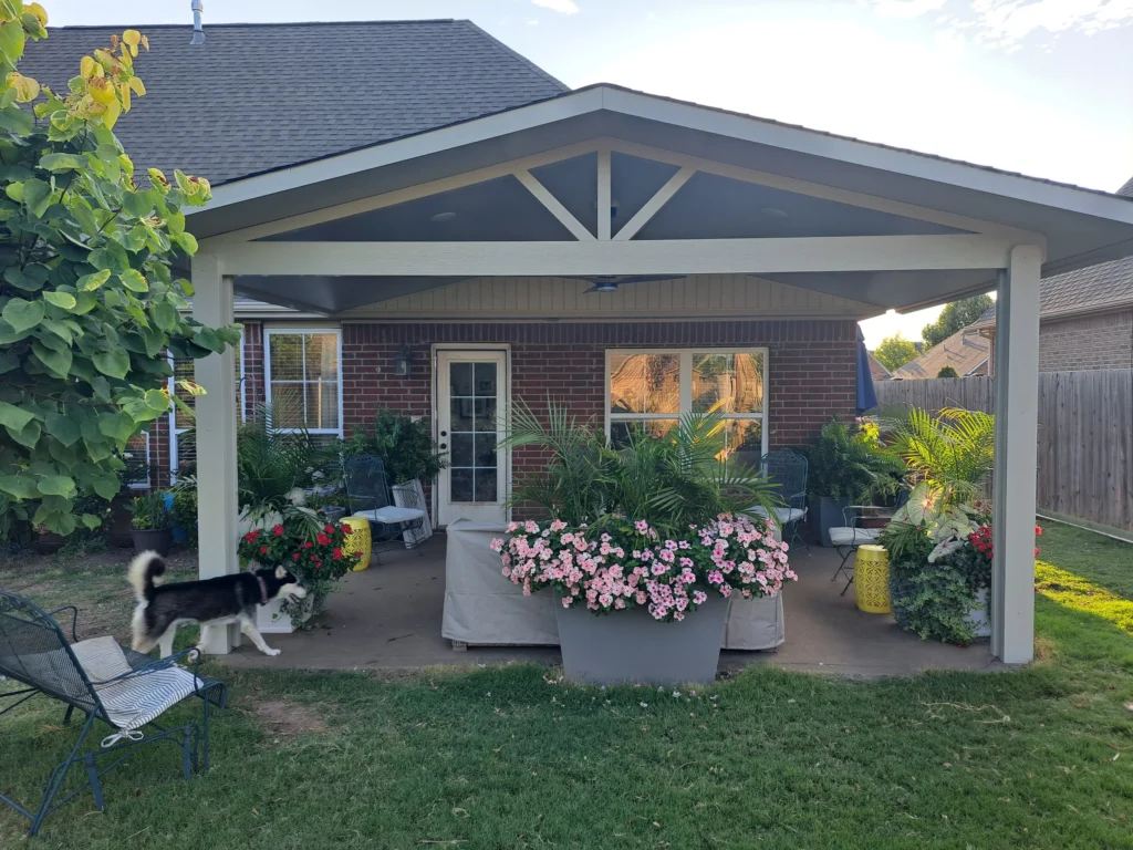 A backyard patio with a covered roof, lush potted plants, outdoor furniture, and a black and white dog walking on the grass near the patio. The house is brick with large windows and a wooden fence in the background.