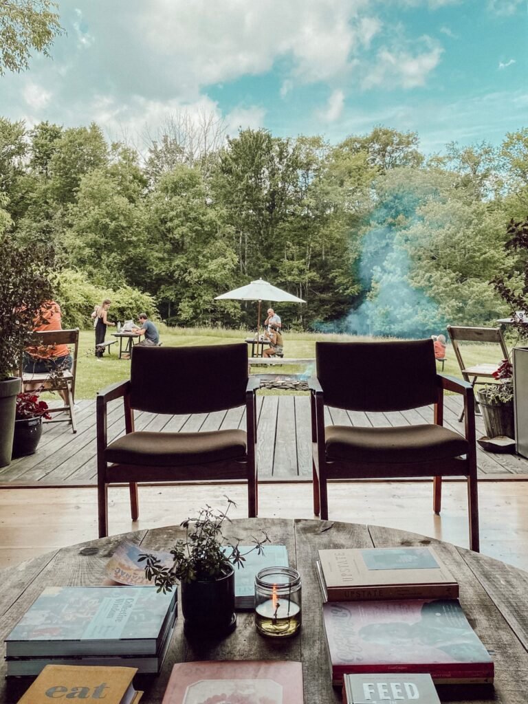 Two empty chairs on a porch face a grassy yard, where people sit and stand near a smoky firepit. Trees and a cloudy sky are in the background. Books and a small plant rest on a table in the foreground.