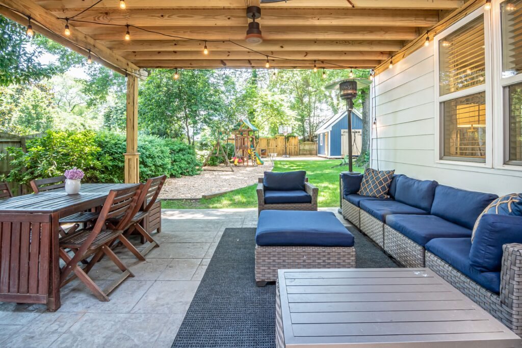 Covered patio with string lights, wooden dining set, wicker sofa and chairs with blue cushions, outdoor rug, and a backyard with grass, trees, a playground, and a garage in the background.