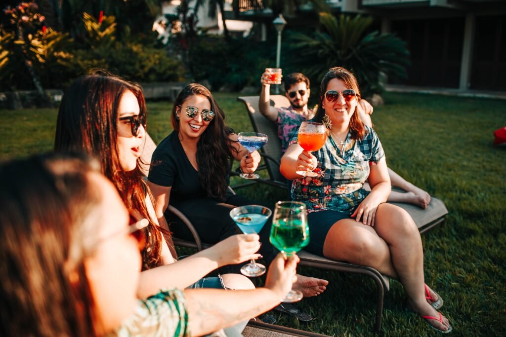 Five people sit on lounge chairs outdoors, smiling and holding colorful cocktails. They appear to be enjoying a sunny day together on a grassy lawn with tropical plants in the background.