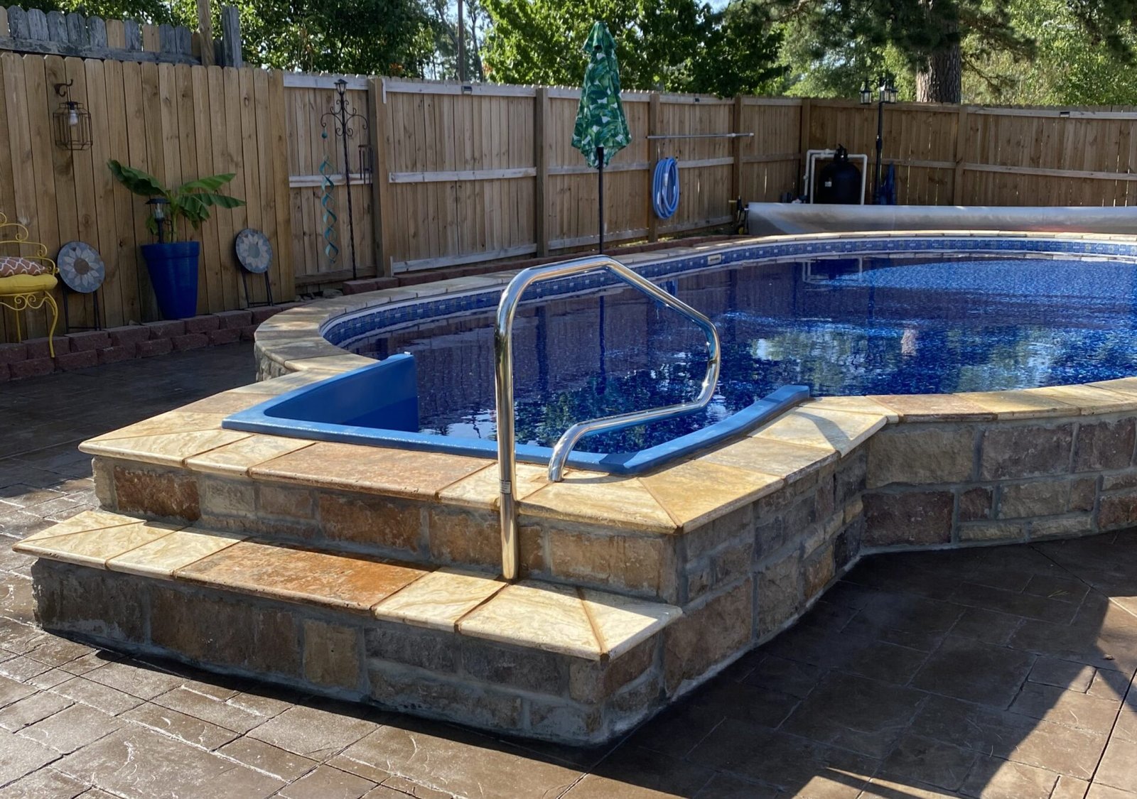 A backyard swimming pool with a stone tile edge, metal handrail, and steps leads into the water. The pool is surrounded by a wooden fence, lush trees, and patio furniture under a sunny sky for relaxing poolside moments.